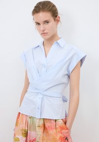 Vaaleansininen, raidallinen kietaisupaita lyhyillä hihoilla, kauluksella ja vyötäröllä solmimiseksia. Yhdistetty kukkakuvioiseen, moniväriseen hameeseen.