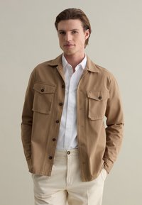 Giacca marrone chiaro con due tasche sul petto e chiusura a bottoni. Realizzata in cotone con una texture liscia, indossata sopra una camicia bianca.