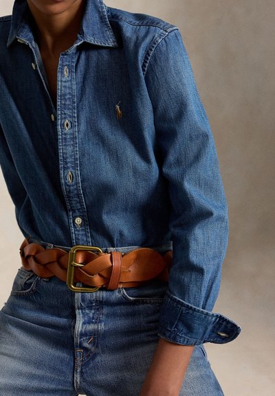 Persoon draagt een blauw denim overhemd en jeans met een brede gevlochten bruine leren riem met een messing gesp.