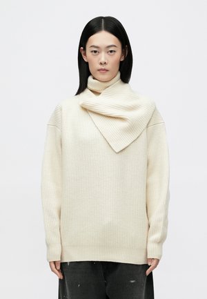 Un pull en tricot couleur crème avec un col montant et un panneau avant superposé, présentant une texture côtelée et des manches longues.