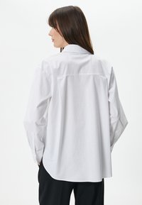 Camicia bianca a maniche lunghe con colletto classico, cucitura della spalla posteriore e polsini. Realizzata in tessuto leggero, presenta un orlo curvo e una vestibilità comoda.