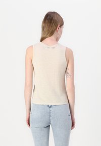 Beige ermeløs topp laget av mykt stoff, med rund halsutskjæring og en subtil merkevarelogo bak. Bakvindu med lyseblå jeans.