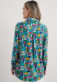 Long-sleeve shirt in turquoise with a geometric pattern of circles and stripes in pink, yellow, green, and blue. Features a collar and button cuff.