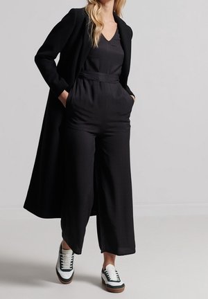 Femme portant une combinaison noire à jambes larges, un long manteau noir et des baskets blanches avec des détails noirs, debout les mains dans les poches.