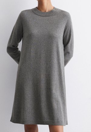 Woman wearing a long-sleeve, knee-length, grey knit sweater dress against a plain white background.