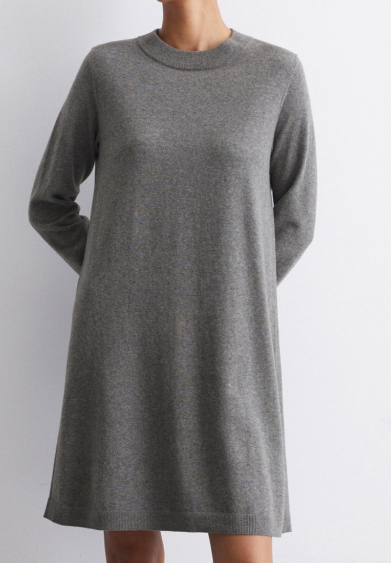 Femme portant une robe pull en maille grise, manches longues, longueur genou, sur un fond blanc uni.