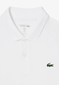 Vit polo med andningsbart tyg, slim fit, tre knappar och en grön Lacoste-logotyp på vänster bröst. Texturerade axelpaneler.