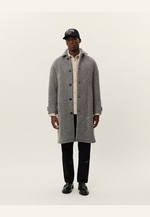 Gray herringbone coat with a collar, front buttons, and side pockets, worn over a light shirt and black pants, paired with black shoes.