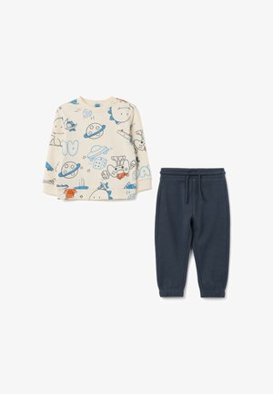 Maglietta a maniche lunghe color beige chiaro con stampe di cartoni animati, abbinata a pantaloni sportivi a gamba affusolata blu scuro con vita elasticizzata.