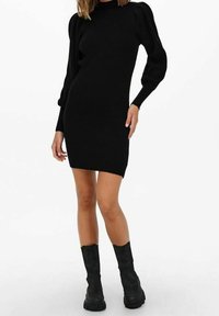 Robe noire côtelée avec un col montant, des manches longues bouffantes et une silhouette ajustée. Portée avec des bottines noires. Texture lisse.