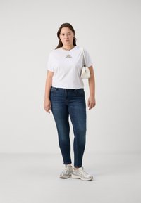 White short-sleeve t-shirt with a sun graphic, dark blue skinny jeans, and light gray sneakers. Model holds a small white handbag.
