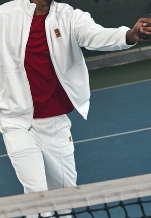 Biały sportowy dres z czerwoną koszulką pod spodem. Kurtka na zamek błyskawiczny z małym logo. Zawodnik gotowy z rakietą na korcie tenisowym.