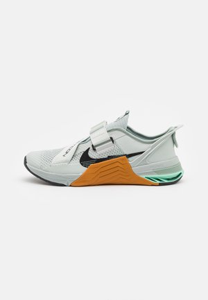 Jasnoszary but sportowy z czarnym logo Nike, pomarańczowym wsparciem śródpodeszwy, miętowym detalem na pięcie i dwoma szerokimi paskami na górze.