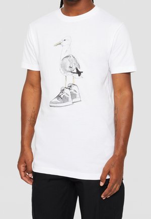 Homme portant un t-shirt blanc avec un graphisme d'une mouette posée sur une paire de baskets, associé à un pantalon noir.
