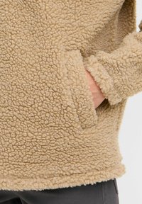 Pull-over en polaire beige avec une surface texturée et pelucheuse. Il dispose d'une poche kangourou et de manches à poignets. L'ourlet est légèrement élastiqué.