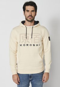 Sudadera color crema con cordones, bolsillo frontal y el texto "KOROSHI" en negro. Tejido suave y puños acanalados. Corte casual.