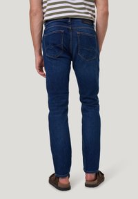 Donkerblauwe denim jeans met een slim fit, met twee achterzakken en subtiele stikseldetails. G gedragen met bruine instapschoenen.
