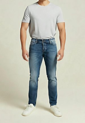 Mężczyzna ubrany w jasnoszary t-shirt, niebieskie obcisłe jeansy i białe trampki stoi na jednolitym tle.