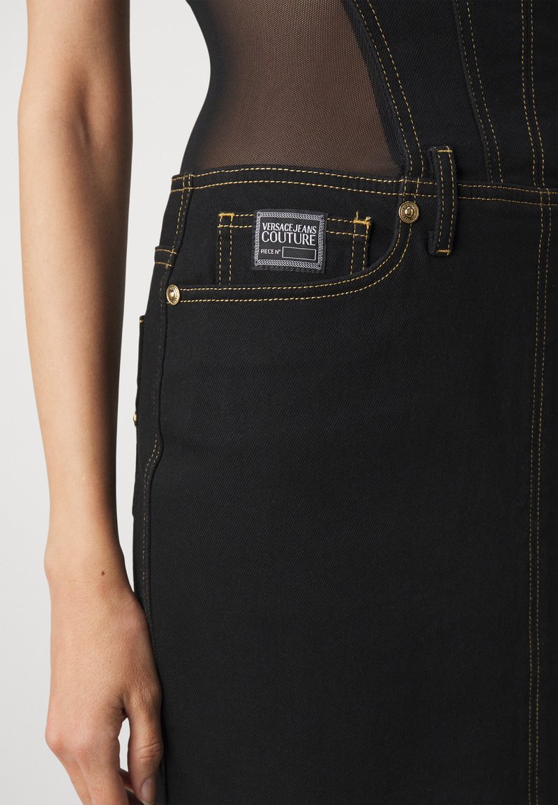 Jean en denim noir avec des coutures jaunes, dotés d'une étiquette de logo sur la poche arrière et de matériel en métal visible. Texture lisse et design ajusté.