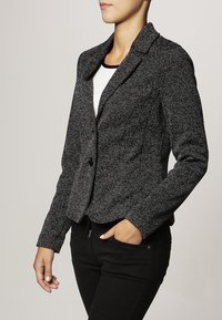 Blazer texturé noir et blanc avec col cranté, fermeture à un seul bouton et coupe ajustée, associé à un haut blanc et un pantalon noir.