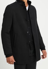 Cappotto nero con colletto alto, tessuto strutturato, tre bottoni e tasche laterali. Abbinato con un look a strati sottostante.