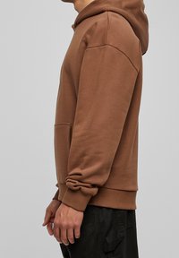 Brun huvtröja med en framficka, ribbade ärmmuddar och nederkant, tillverkad av ett mjukt tyg. Har en dragsko och en avslappnad passform.