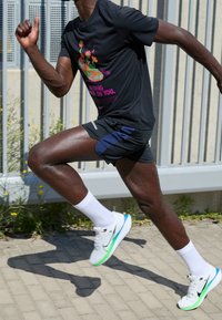 Černé sportovní tričko s barevným květinovým potiskem, tmavě modré šortky a bílé tenisky s zelenými a modrými akcenty, které nosí osoba při běhu.