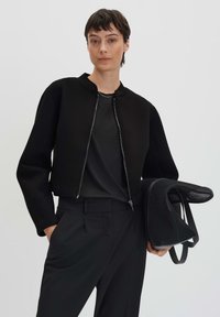 Veste noire courte avec un col rond et des détails de fermeture éclair, associée à une chemise noire et un pantalon. Tenant un sac en maille noir.
