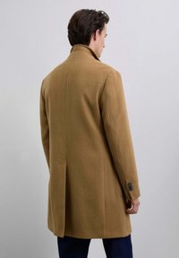 Hombre vestido con un abrigo de camel de longitud a la rodilla, con un cuello alzado y botones oscuros en los puños, de pie contra un fondo claro y liso.