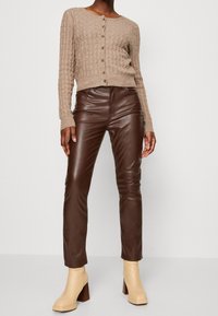 Pantalones de cuero marrón y un cárdigan beige acanalado con cierre de botones, que presenta un escote redondo y mangas largas ajustadas. Botines de tacón beige.