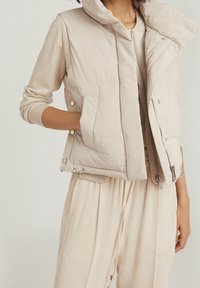Beżowa pikowana kamizelka z kapturem, wyposażona w przedni zamek błyskawiczny i zatrzaski, uzupełniona bocznymi kieszeniami oraz regulowanym ściągaczem u dołu.