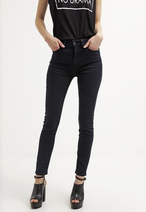 Femme portant un jean skinny noir, un haut sans manches noir avec l'inscription « NO DRAMA » et des chaussures noires à talons hauts ouvertes à l'avant avec bride à la cheville, debout les mains dans les poches.