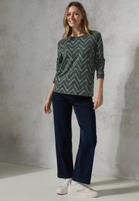 Chemise à manches longues verte à motifs avec un design en zigzag, associée à un jean ample bleu foncé et des baskets blanches sur un fond clair.