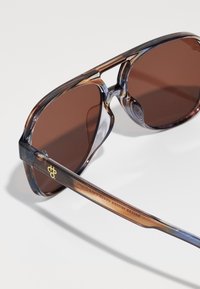 Ochelari de soare cu nuanțe de marron, cu un cadru translucid brown și albastru, având un design tăiat unic și un logo pe braț.