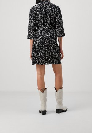 Femme portant une robe noire avec des motifs de visage en ligne continue blanche, des bottes de cowboy blanches mi-mollet, debout devant un fond uni.