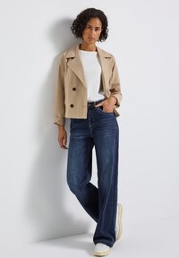 Manteau trench court beige avec double boutonnage, associé à un jean large taille haute bleu foncé et des baskets blanches. Textures de tissu lisses.