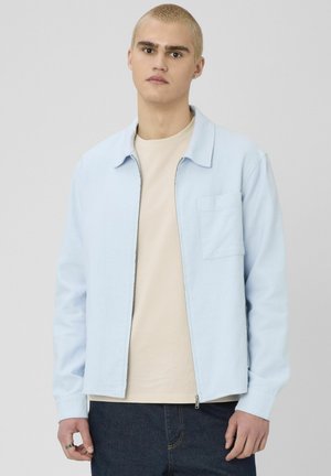 Jeune homme aux cheveux blonds courts portant une veste zippée bleu clair sur un t-shirt beige et un jean foncé, debout devant un fond gris.