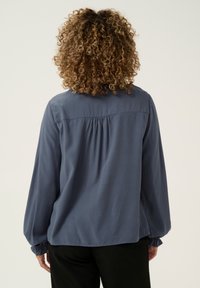Blusa a maniche lunghe di colore blu scuro, con una texture liscia, dettagli arricciati sul retro e polsini elasticizzati. Abbinata a pantaloni neri.