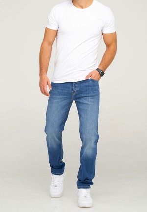 Mann trägt ein schlichtes weißes T-Shirt, blaue Jeans, weiße Turnschuhe und eine schwarze Smartwatch, steht mit einer Hand in der Tasche vor hellem Hintergrund.