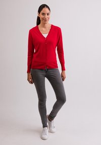 Röd cardigan med v-hals och knappstängning, kombinerad med grå slim-fit jeans och vita sneakers. Slät textur och figurnära design.