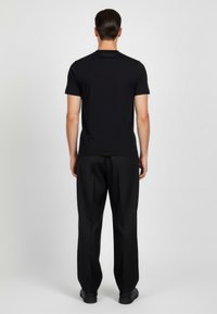 Camiseta negra de manga corta con cuello redondo, hecha de una tela suave. Presenta un sutil detalle del logo en la parte superior de la espalda. Combinada con pantalones negros.