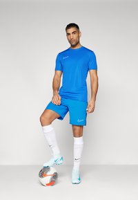Blå idretts skjorte og Shorts laget av lette materialer, kombinert med hvite sokker og blå fotballsko. Individuell poserer med en fotball.