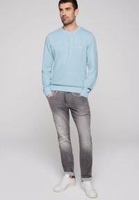 Sudadera azul claro con texto en relieve "CAMP DAVID", combinada con jeans de denim gris y zapatillas blancas. Tejido suave y corte casual.