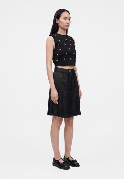 Femme portant un crop top noir sans manches avec des œillets dorés, une jupe plissée noire longueur genou et des chaussures Mary Jane noires, debout de côté.