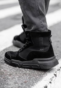 Personne portant des baskets montantes noires et un pantalon gris, debout sur l'asphalte près d'une ligne blanche peinte au sol.