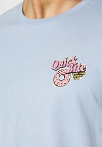 Světle modré bavlněné tričko s potiskem v růžové a žluté barvě, na kterém je design donutu a text: "Klub milovníků rychlého občerstvení".