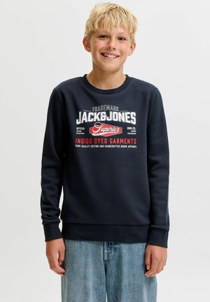 Blond dreng smilende iført en sort Jack & Jones sweatshirt med hvid og rød tekst og lyseblå jeans, stående foran en ensfarvet baggrund.