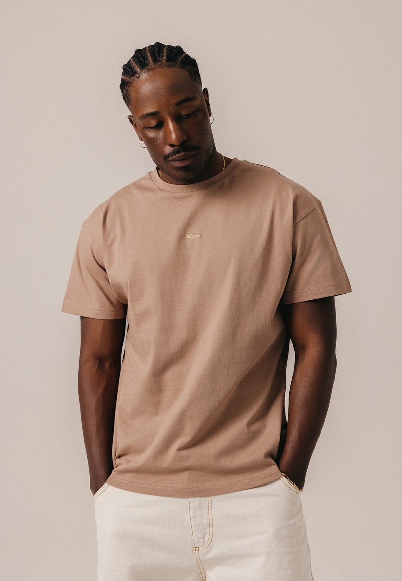 Homme aux cheveux tressés portant un t-shirt beige et un pantalon blanc, debout devant un fond clair uni, les mains dans les poches.