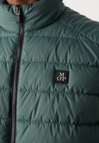 Grüne, gepolsterte Jacke mit gesteppter Optik, hohem Kragen und schwarzem Reißverschluss. Mit einem quadratischen Logo-Patch mit „M O’P“ auf der Brust.