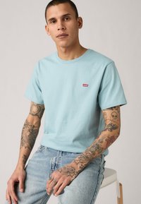 Tricou din bumbac bleu deschis cu guler rotund, av�ând un mic logo Levi's roșu pe piept, purtat cu blugi deschiși.